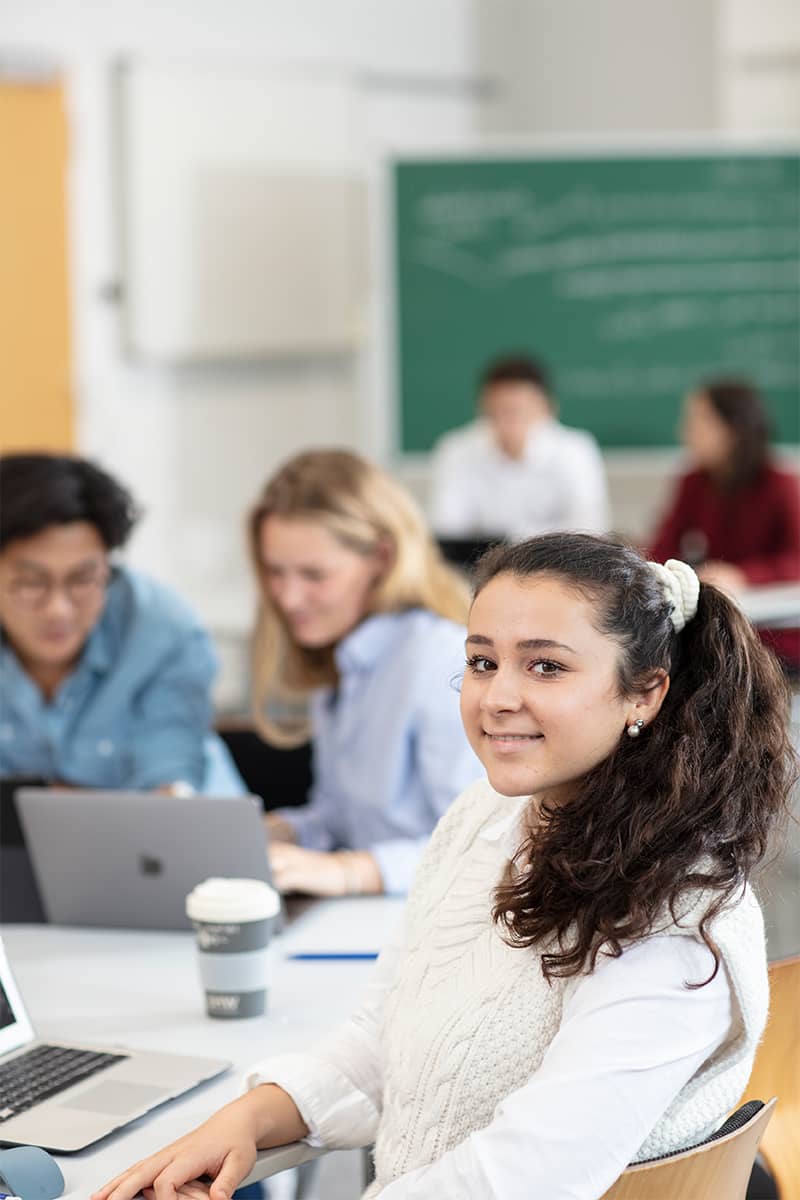 Lächelnde Studentin im Vordergrund. Im Hintergrund lernen zwei Studierende. 