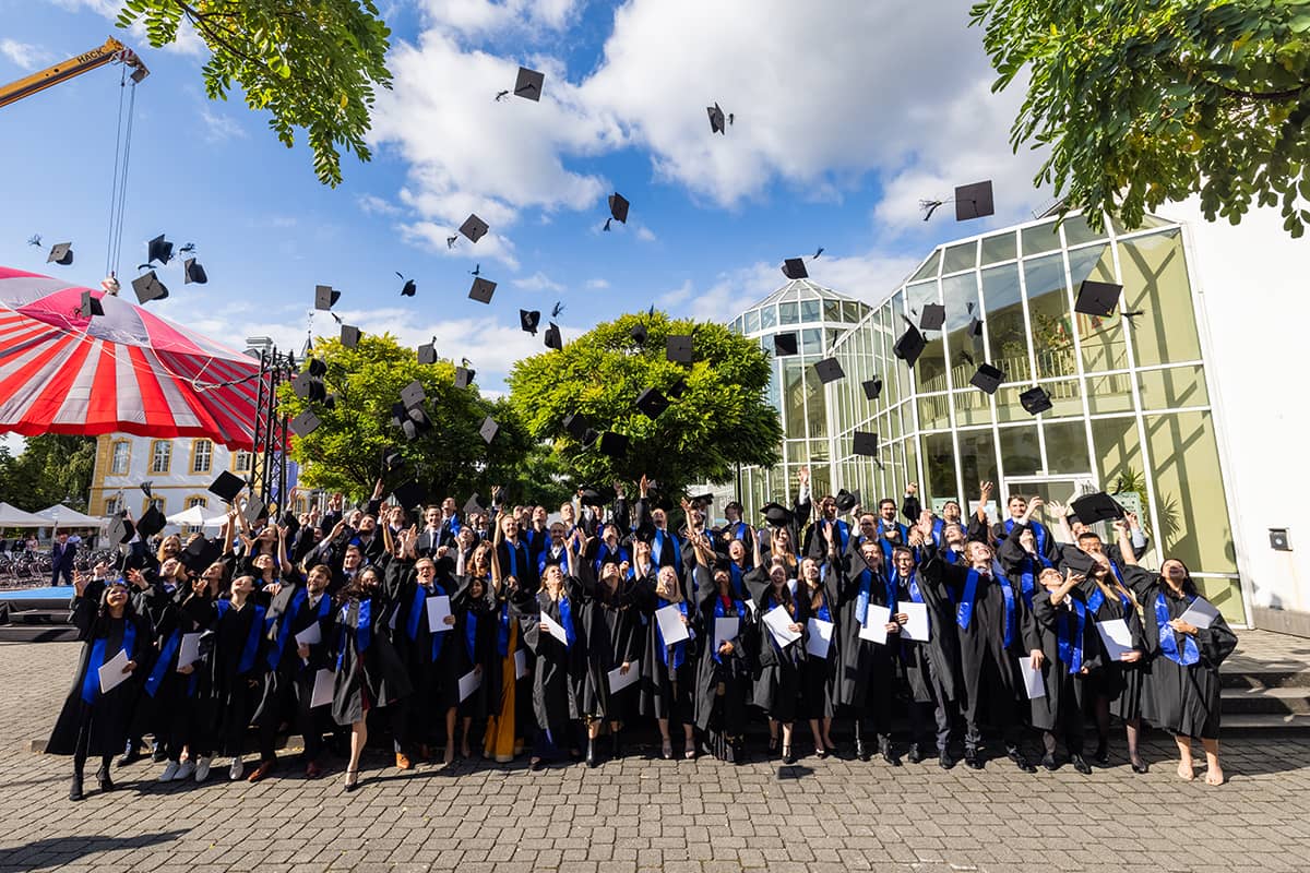 A group photo of the Master graduates throwing their caps into the air on the Burgplatz
