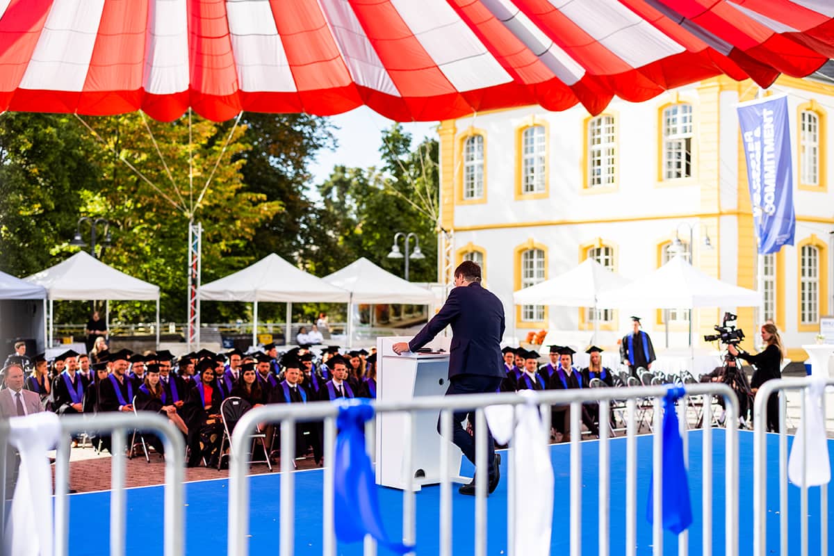 Dean Professor Markus Rudolf addresses the seated guests from the podium on the Burgplatz at the WHU Graduation Day