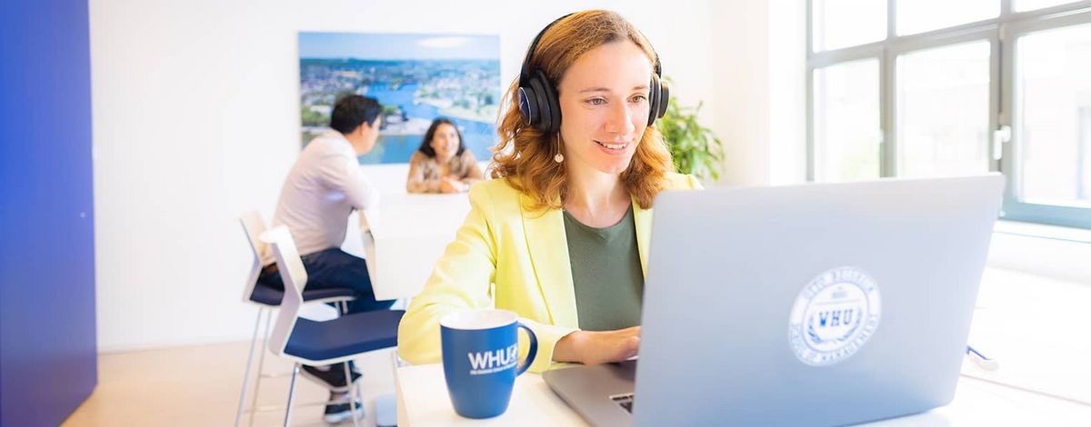 [Translate to English:] A female MBA student wearing a yellow jacket and headphones works at her notebook