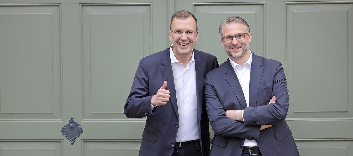 Utz Schäffer and Marko Reimer stand in front of a door on the WHU Vallendar campus and smile into the camera